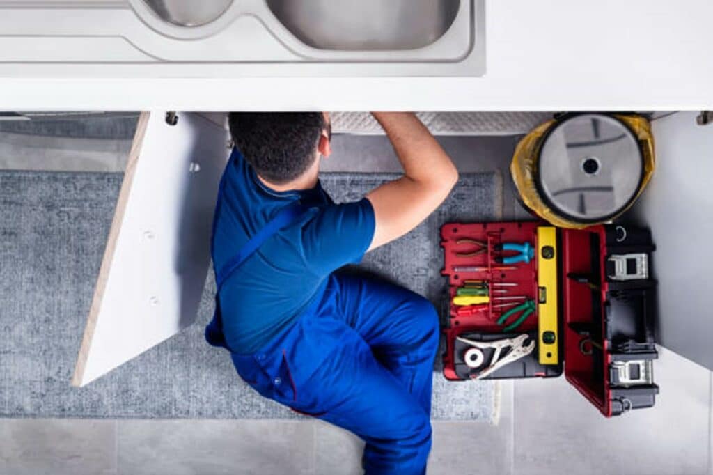 A plumber with a tool kit leans under a sink to identify the cause of blocked drains